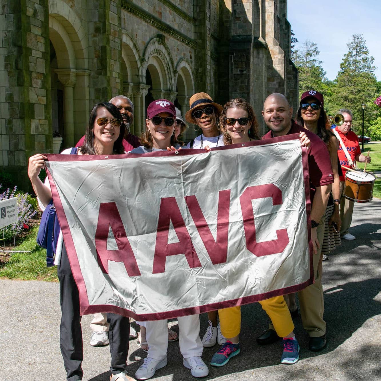 A group of people holding a large grey banner with the letters AAVC on it are smiling at the viewer.